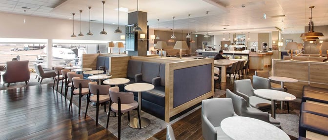 The lounge at London Heathrow Airport features booth and table seating with views of the runway and a bar area.