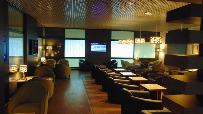 The Sarajevo International Airport business lounge features comfortable seating with individual tables and a flight information display screen.