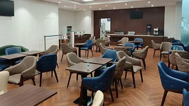 The seating area at The Lounge Terminal 2 Sardar Vallabhbhai Patel International Airport features tables and chairs with a view of the food service counter.