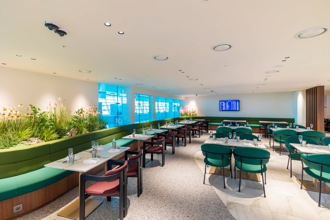 The dining area at MATINA Lounge East, Gate 11, features table seating with greenery and a flight information display.