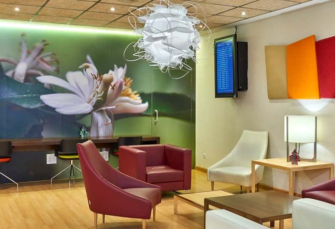 The Sala VIP Azahar at Seville Airport features comfortable seating, a wall-mounted flight information screen, and workstations with charging outlets.