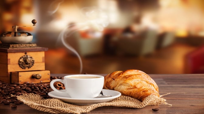 A coffee and croissant are placed on a wooden table, with a manual coffee grinder and scattered beans nearby.