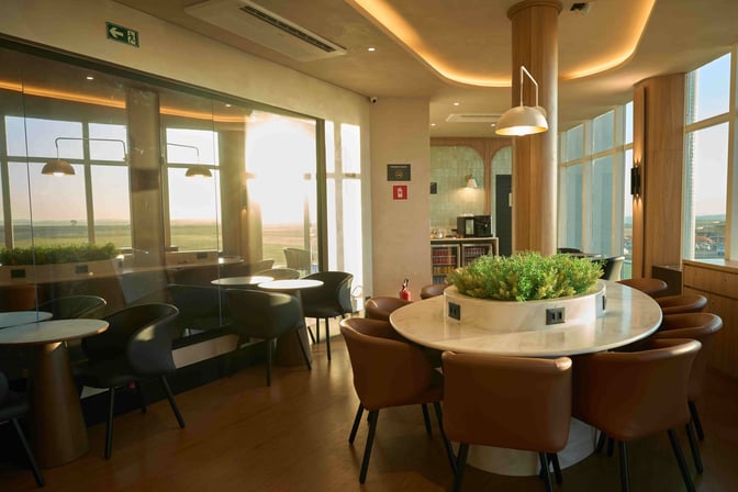 The W Café Lounge at Sílvio Name Júnior Regional Airport features round tables with seating and a central plant arrangement.