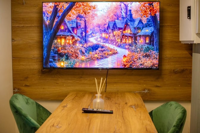A wooden table with green chairs faces a wall-mounted TV displaying a scenic image, accompanied by a remote and diffuser.