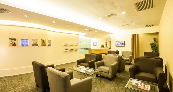 The Sky Lounge at Sultan Abdul Aziz Shah Airport features comfortable armchairs and glass tables, with a television and decorative wall art.
