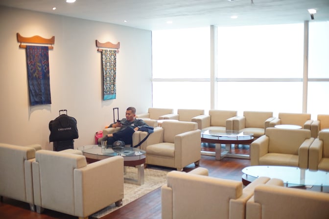 The seating area in Concordia Lounge at Sultan Hasanuddin International Airport features multiple armchairs and glass-topped tables for passenger relaxation.