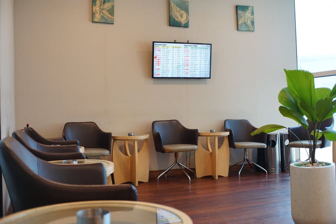The seating area in Concordia Lounge at Sultan Hasanuddin International Airport features comfortable chairs, small tables, and a flight information screen.