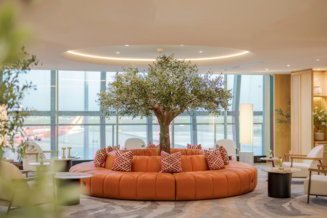 Circular seating surrounds a central decorative tree in The Coral Finest Business Class Lounge Cocoon at Suvarnabhumi Airport, offering airport views.