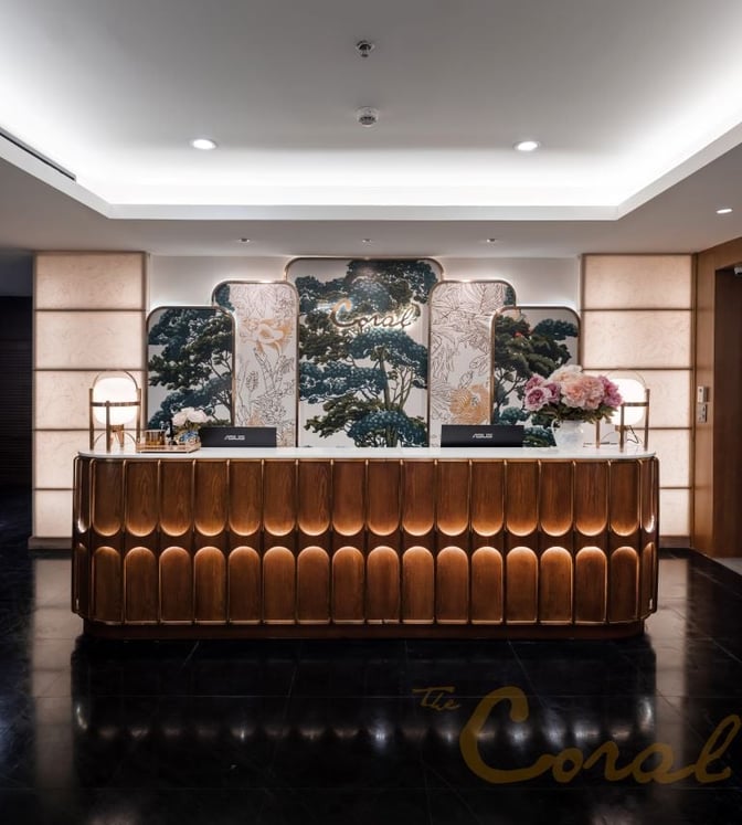The reception desk at The Coral First Class Lounge in Suvarnabhumi Airport features check-in counters and decorative floral arrangements.