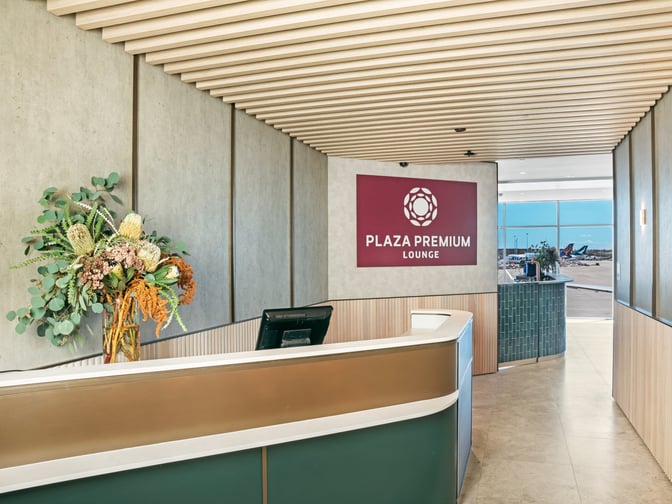 The Plaza Premium Lounge entrance at Sydney Airport features a reception desk and a sign, with views of the tarmac beyond.
