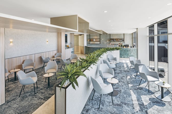 The Plaza Premium Lounge at Sydney Airport features a seating area with armchairs and small tables, adjacent to a bar.