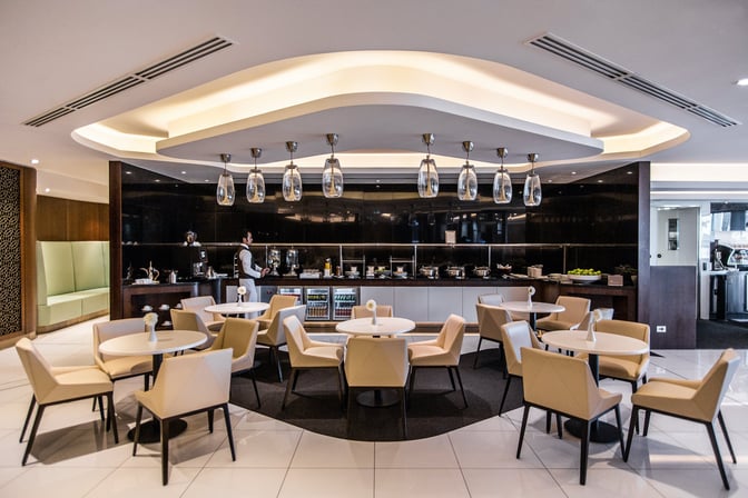 The dining area at The House lounge in Sydney Airport features round tables, seating, and a buffet with various food options.