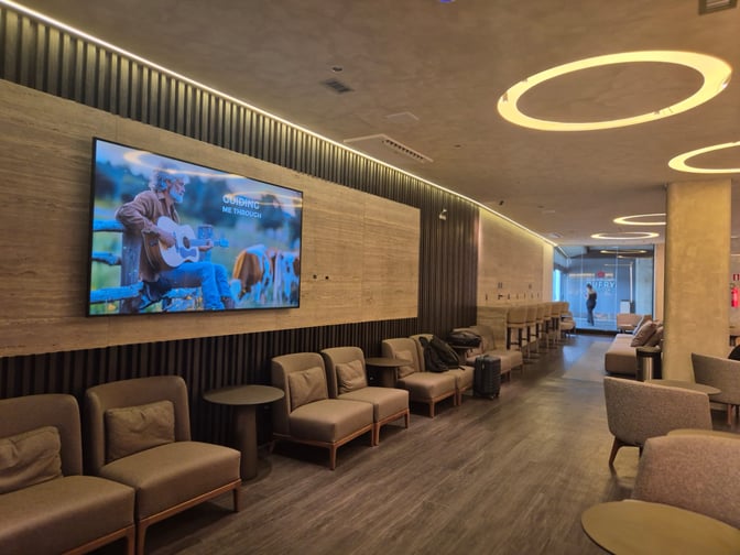 The BRT Lounge at Tancredo Neves International Airport features comfortable seating with a large wall-mounted screen and a bar area.