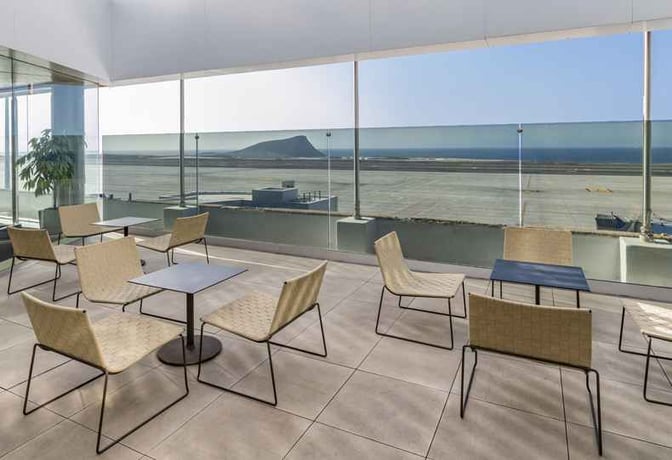 The seating area in Sala Montaña Roja at Tenerife South Airport offers tables and chairs with expansive views of the runway.