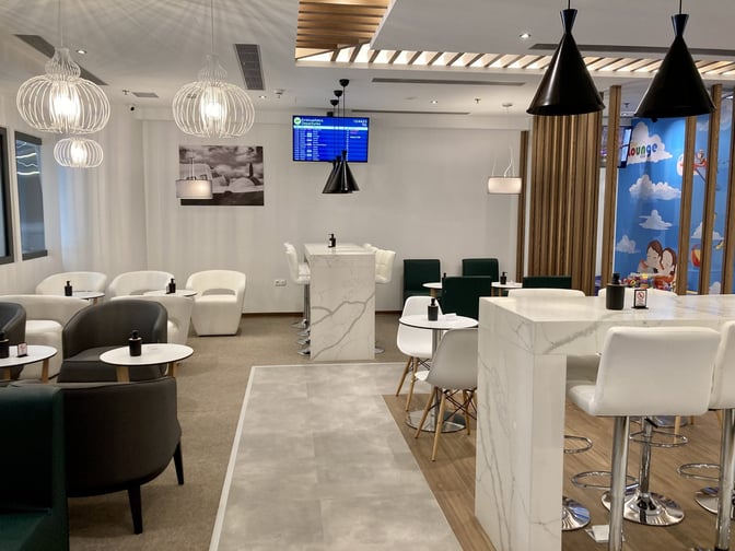 The Lounge Thessaloniki features a seating area with tables and chairs, a bar counter, and a flight information screen.
