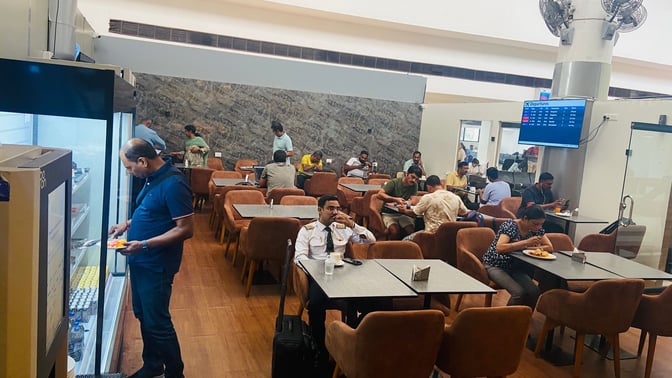 The seating area at Thiruvananthapuram International Airport's domestic lounge features tables and chairs with passengers dining and using electronic devices.
