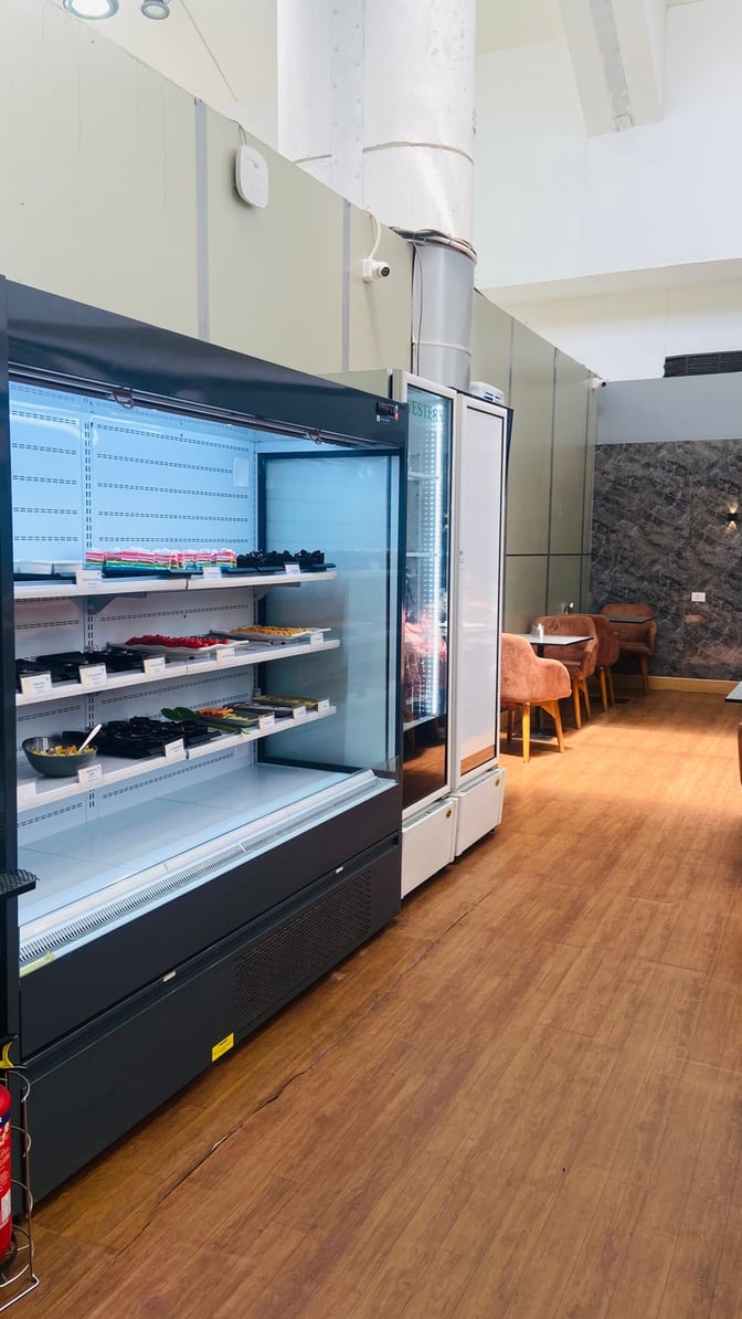 The lounge at Thiruvananthapuram International Airport features a refrigerated display with snacks and a seating area with tables and chairs.