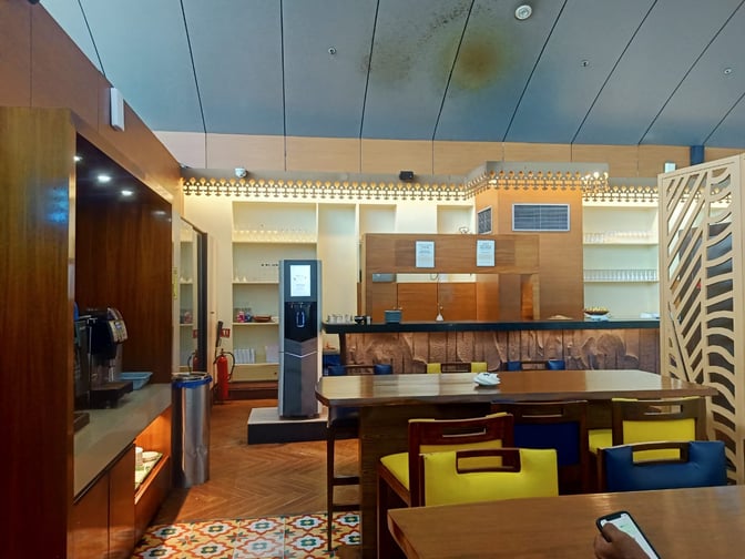 The lounge at Thiruvananthapuram International Airport features a seating area with tables and chairs, adjacent to a self-serve beverage station.