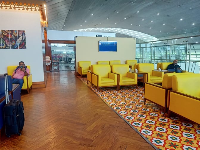 The seating area at Thiruvananthapuram International Airport's lounge features comfortable chairs and tables with airport views and a flight information display.