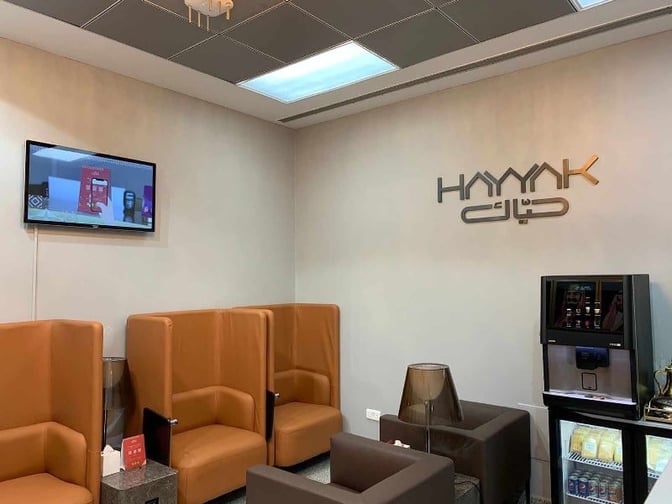 The Hayyak Lounge at Turaif Domestic Airport features high-backed seating, a wall-mounted TV, and a beverage station for refreshments.