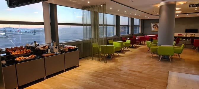 The Austrian Lounge at Vienna International Airport features a seating area with tables and chairs, alongside a self-serve pastry station.