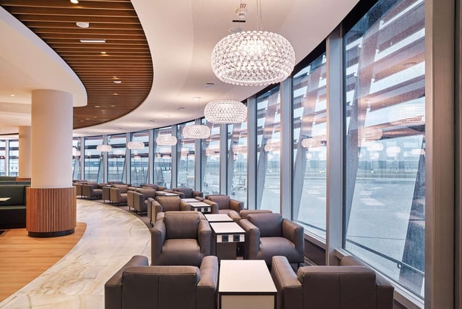 The Vienna Lounge at Vienna International Airport features comfortable seating with tables, offering expansive views of the airport through large windows.